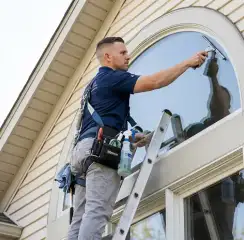 Professional window cleaner on a ladder using a squeegee to clean an exterior arched window, Mesa, AZ.